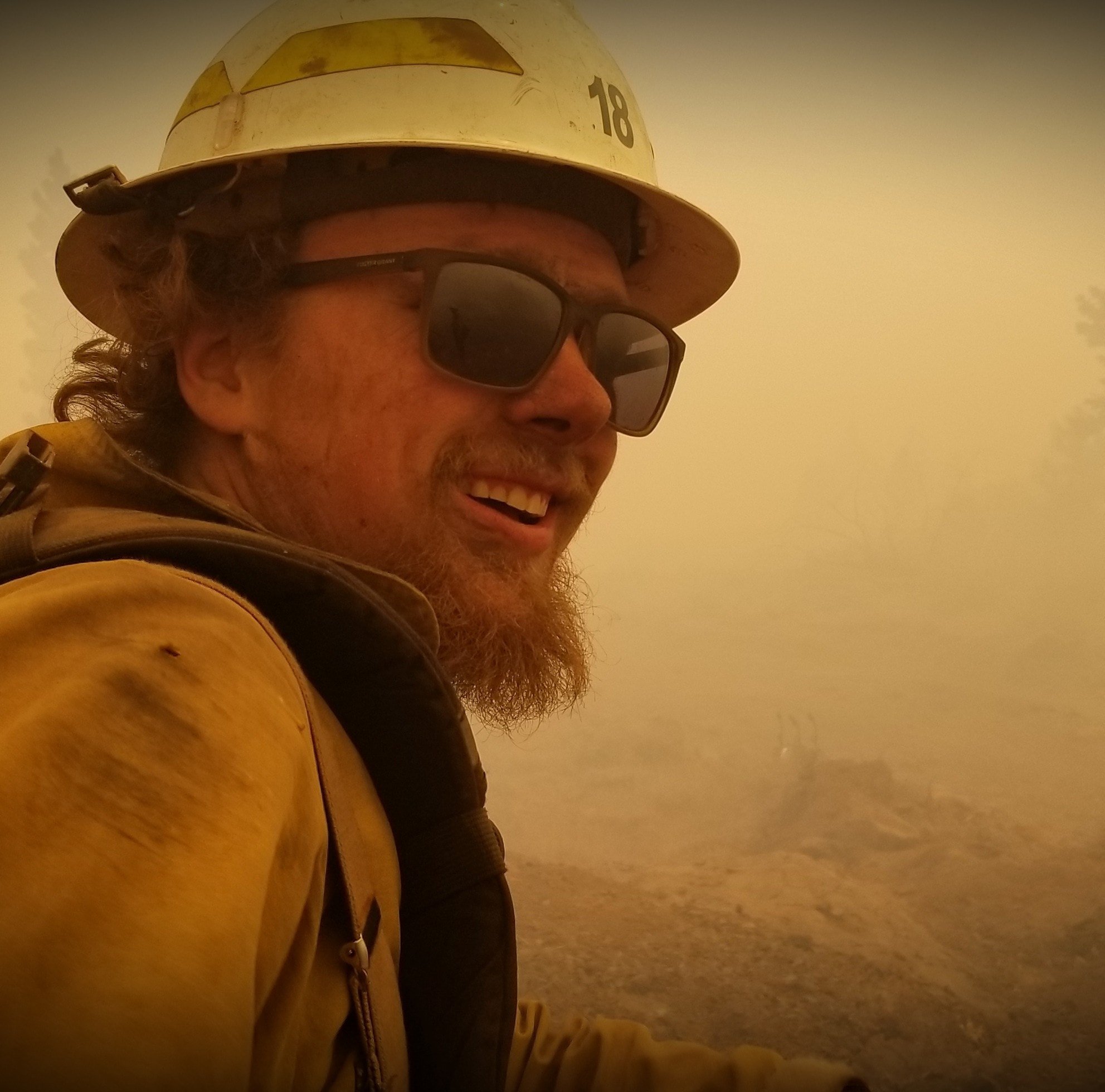 A firefighter wearing a yellow helmet, sunglasses, and a yellow uniform, smiling amidst a smoky background.