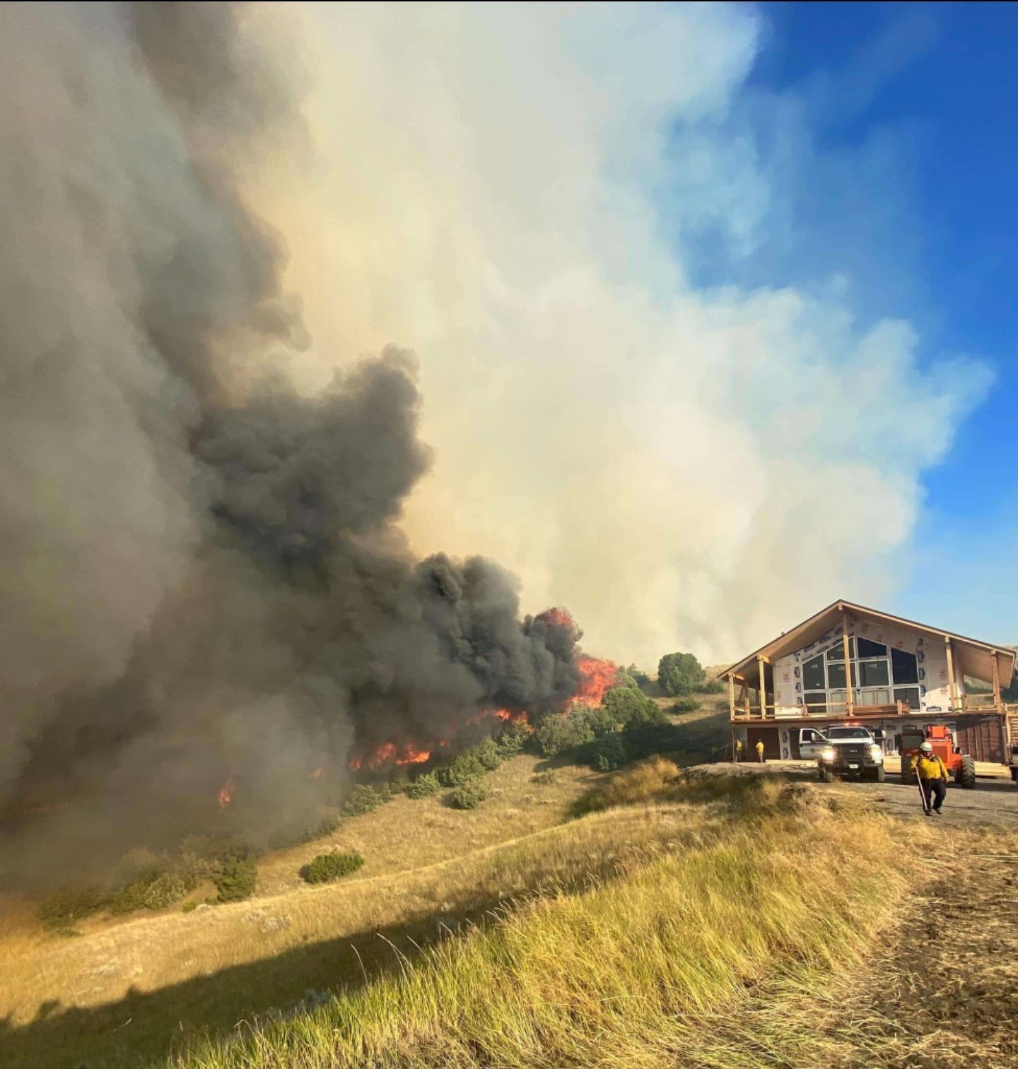 A house on a hillside while a wildfire burns on the hillside, producing thick black smoke and flames.